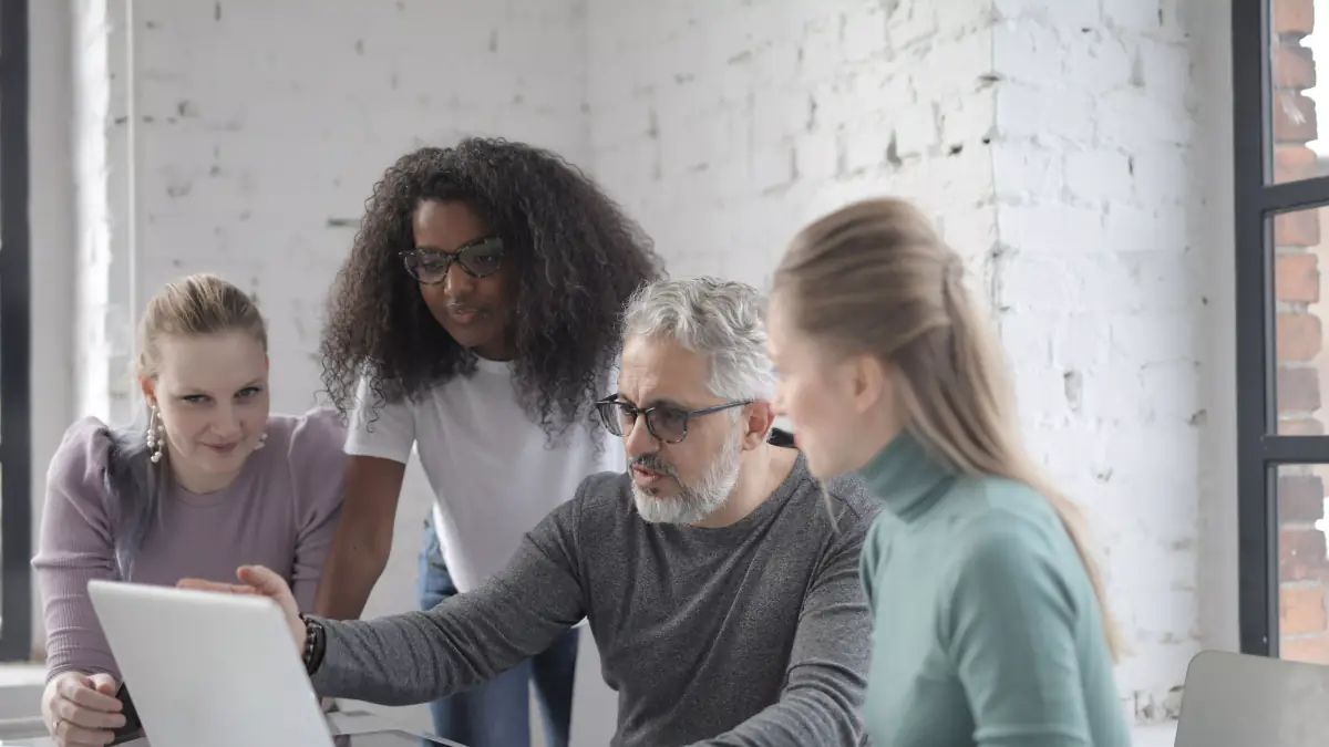 Four colleagues gathered around a laptop, one pointing at the screen while explaining a business project