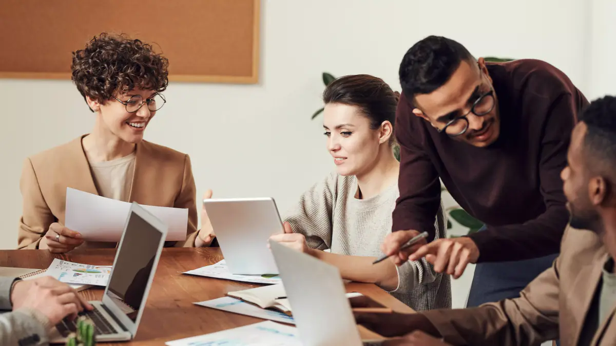 A small group of colleagues at a wooden desk reviewing documents and a tablet together
